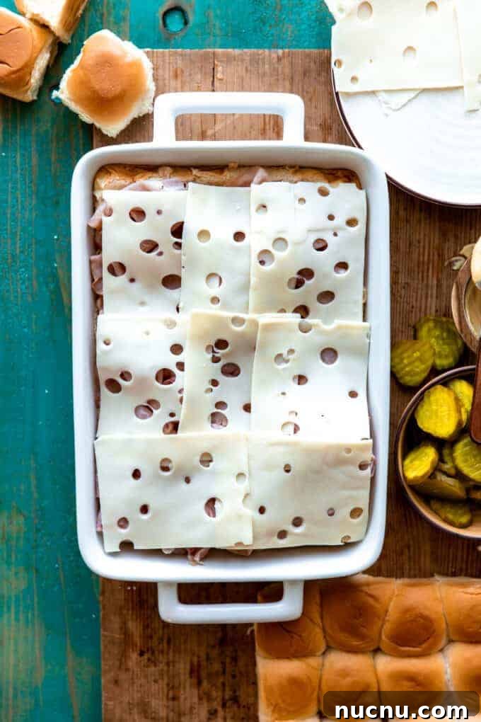 Authentic Cuban Slider Recipe: Mini Cuban Sandwiches to Savor 4 White baking dish showing layers of Hawaiian rolls topped with sliced ham and slices of Swiss cheese.