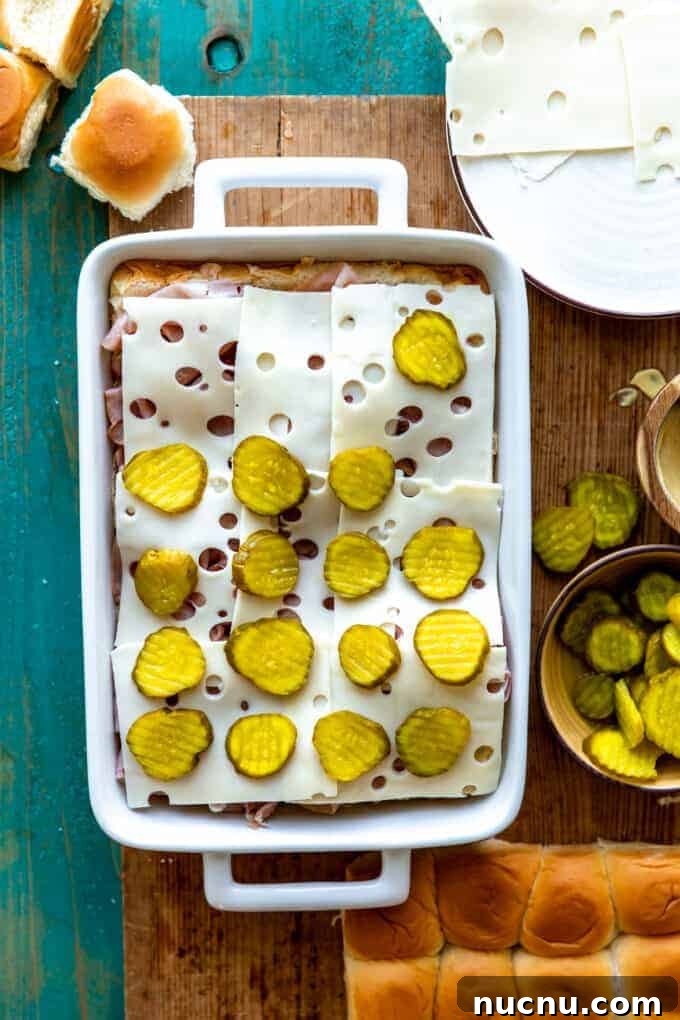 Authentic Cuban Slider Recipe: Mini Cuban Sandwiches to Savor 5 White baking dish with layers of Hawaiian rolls, sliced deli ham, sliced Swiss cheese and pickles.