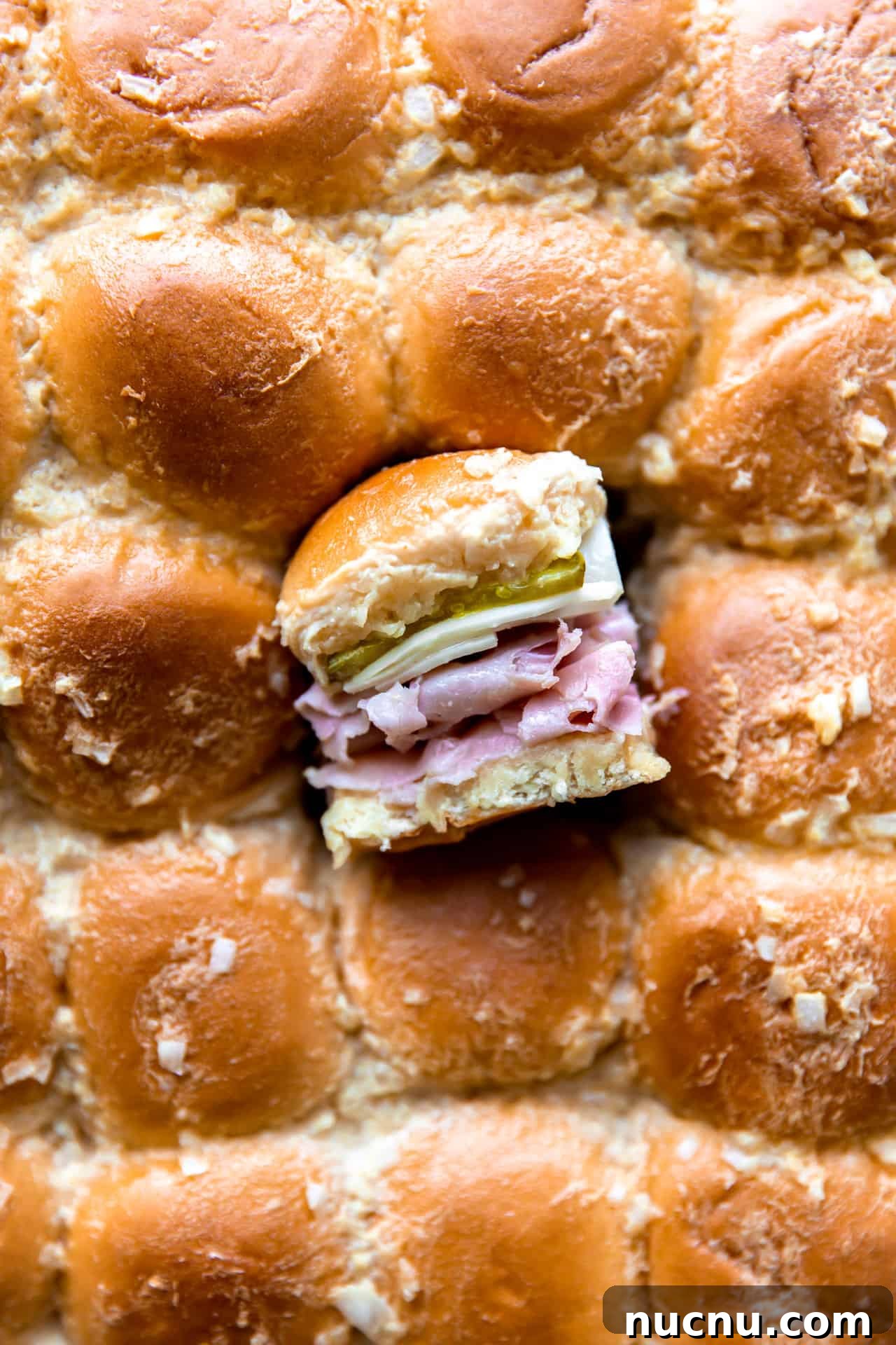 Authentic Cuban Slider Recipe: Mini Cuban Sandwiches to Savor 7 Overhead shot of the tops of sliders with one slider cut out and turned on it's side to show the layers of buns, sliced ham, Swiss cheese and pickles on the inside.