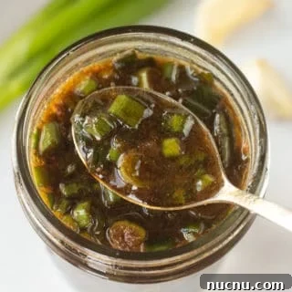 A small spoon dipping into a small mason jar with a brown marinade with chopped green onions in it. Fresh green onions and cloves of garlic are around the jar.