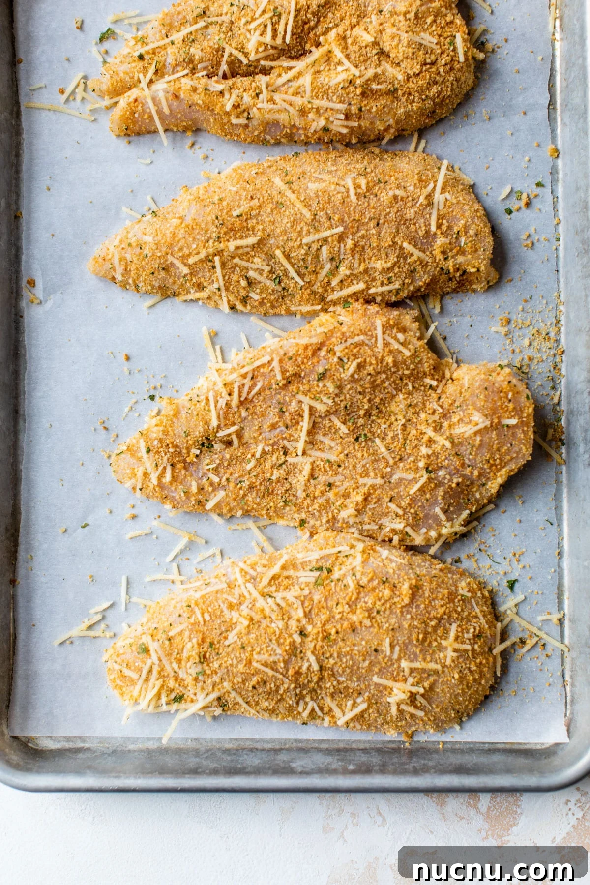 Crispy Baked Parmesan Chicken with Garlic and Herbs 4 raw chicken breasts coated in cheese and breadcrumbs on a baking sheet