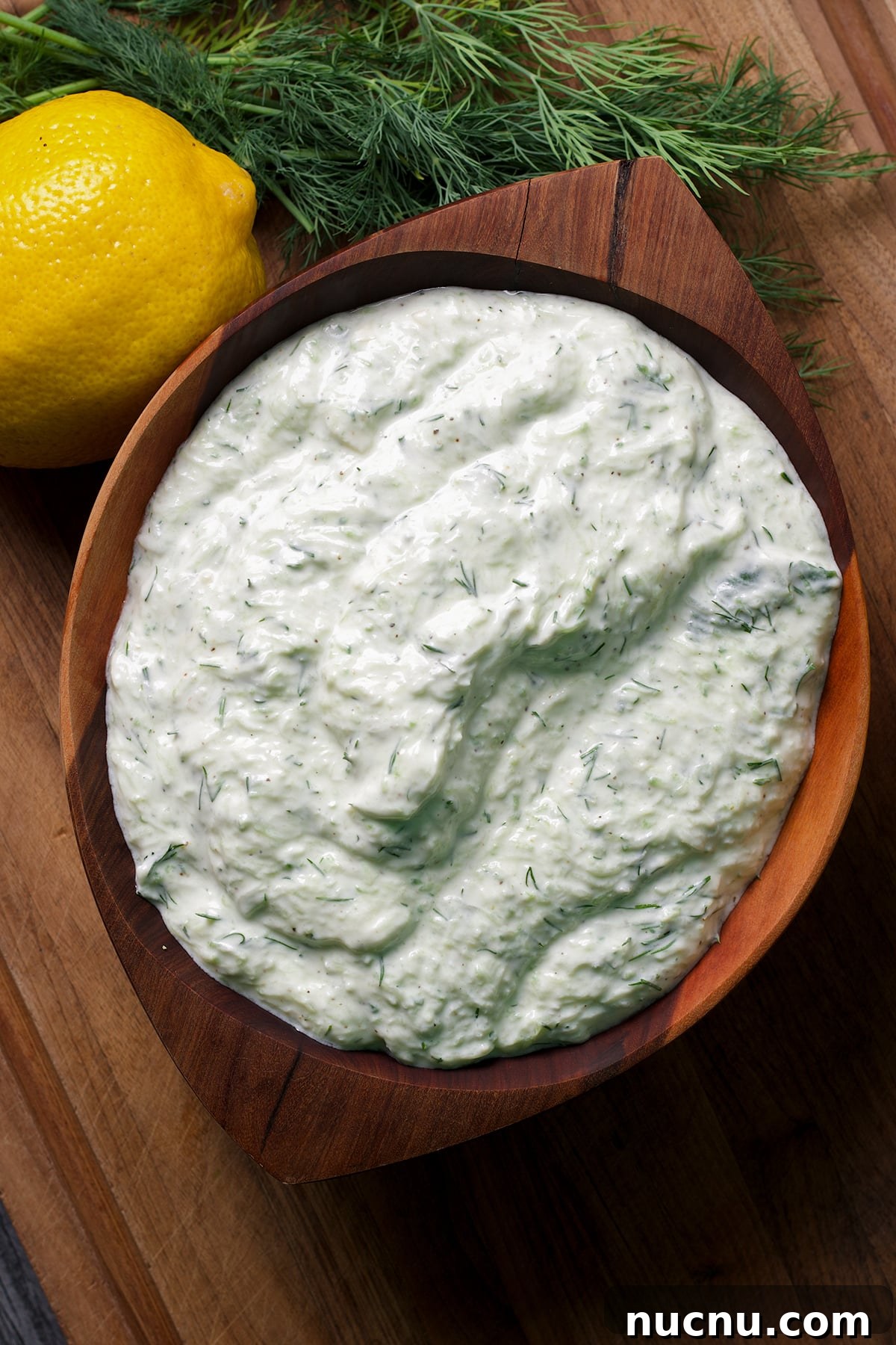 Traditional and Vegan Tzatziki: Authentic Recipes for Both Versions 2 A bowl containing tzatziki sauce rests on a wood cutting board surrounded by fresh dill and lemons.