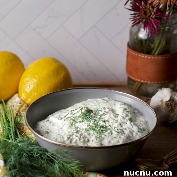 A blue bowl containing vegan tzatziki sauce on a wood board surrounded by fresh dill and lemons.