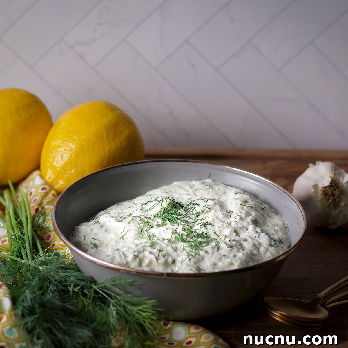 Traditional and Vegan Tzatziki: Authentic Recipes for Both Versions 4 A blue bowl containing vegan tzatziki sauce on a wood board surrounded by fresh dill and lemons.