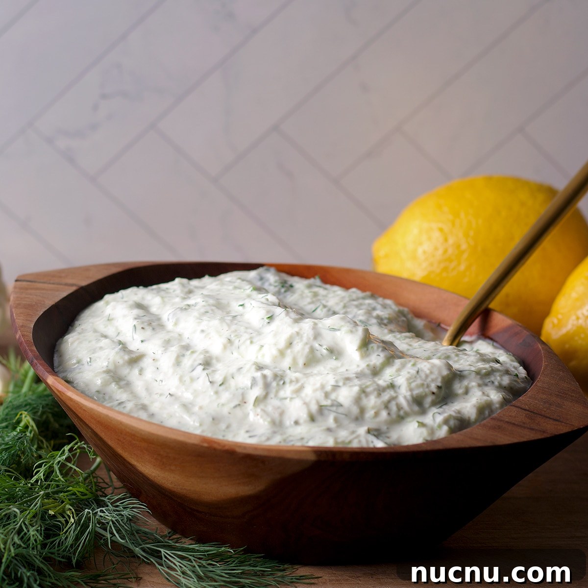 Traditional and Vegan Tzatziki: Authentic Recipes for Both Versions 9 A wood bowl filled with tzatziki sauce with fresh dill and lemons on a cutting board next to the bowl.