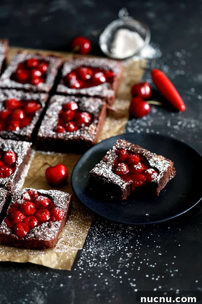 These Fudgy Black Forest Bars definitely have the rich chocolate flavor and moist, dense texture typical of fudge. They’re also the home of puddles of cherry pie filling and studded with dark chocolate chunks before being showered with powdered sugar. The ultimate chocolate and cherry dessert combination. #blackforest #chocolate #cherry #dessertfoodrecipes #desserts Fudgy Black Forest Bars rest on brown parchment on a black background while one bar sits on a black plate.