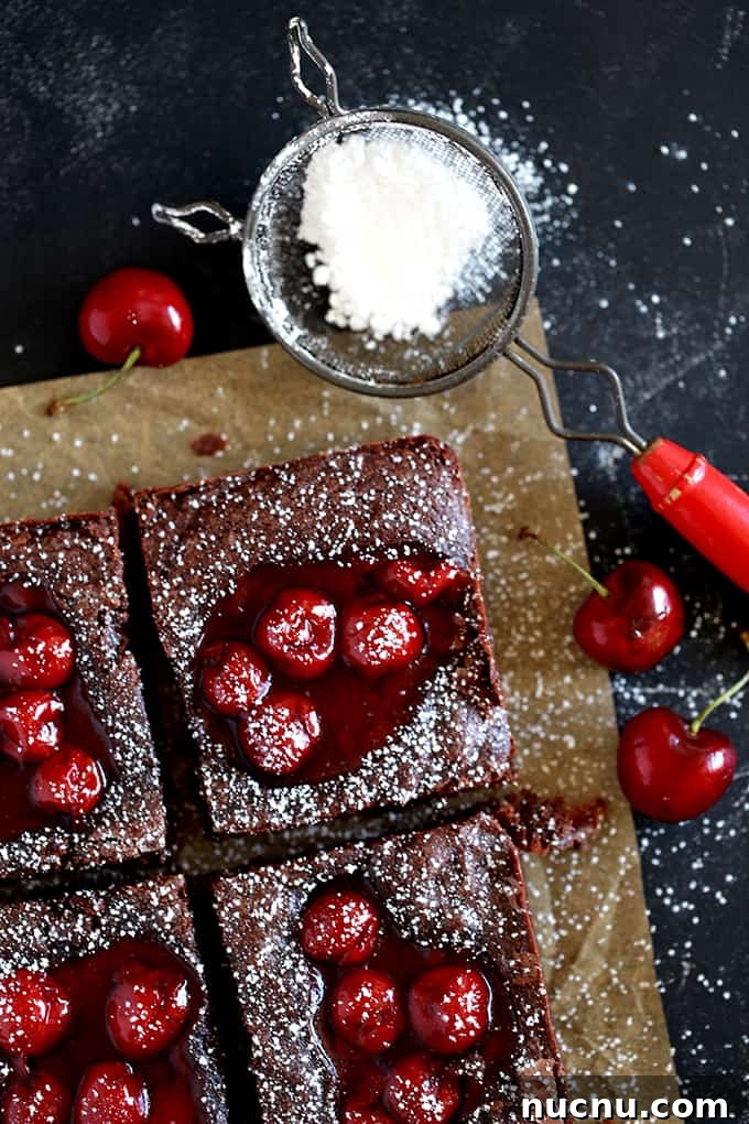 These Fudgy Black Forest Bars definitely have the rich chocolate flavor and moist, dense texture typical of fudge. They’re also the home of puddles of cherry pie filling and studded with dark chocolate chunks before being showered with powdered sugar. The ultimate chocolate and cherry dessert combination. #blackforest #chocolate #cherry #dessertfoodrecipes #desserts Fudgy Black Forest Bars sit on brown parchment on a dark background with powdered sugar in a sifter.