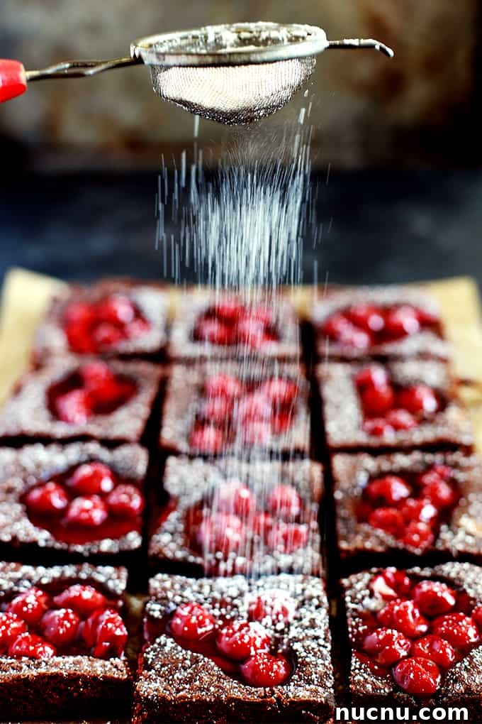 These Fudgy Black Forest Bars definitely have the rich chocolate flavor and moist, dense texture typical of fudge. They’re also the home of puddles of cherry pie filling and studded with dark chocolate chunks before being showered with powdered sugar. The ultimate chocolate and cherry dessert combination. #blackforest #chocolate #cherry #dessertfoodrecipes #desserts Powdered sugar is sifted onto Fudgy Black Forest Bars on brown parchment sitting on a dark background.