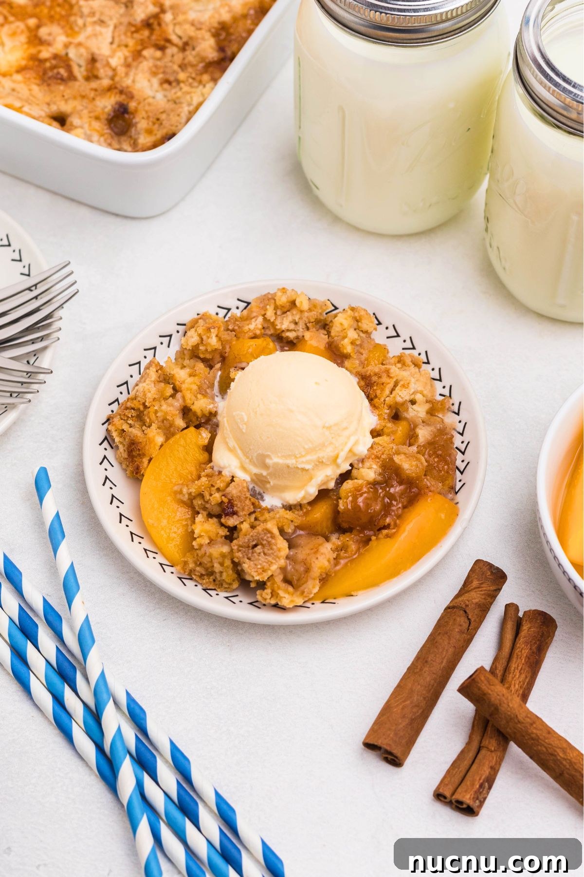 Irresistible Peach Dump Cake Recipe for Quick Summer Desserts 8 Peach cobbler scooped out and served on a small plate, in front of the whole dish, with blue striped straws and cinnamon sticks on the table.