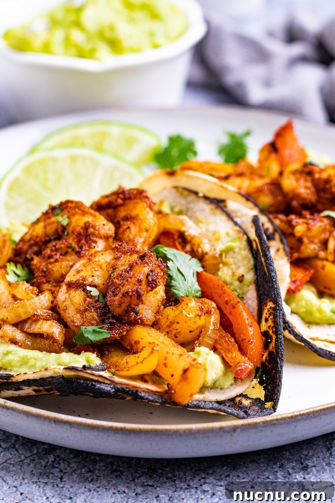 A close up, angled shot of sheet pan shrimp fajitas. Shrimp and peppers are in a tortilla, on a plate, guacamole is in the background.