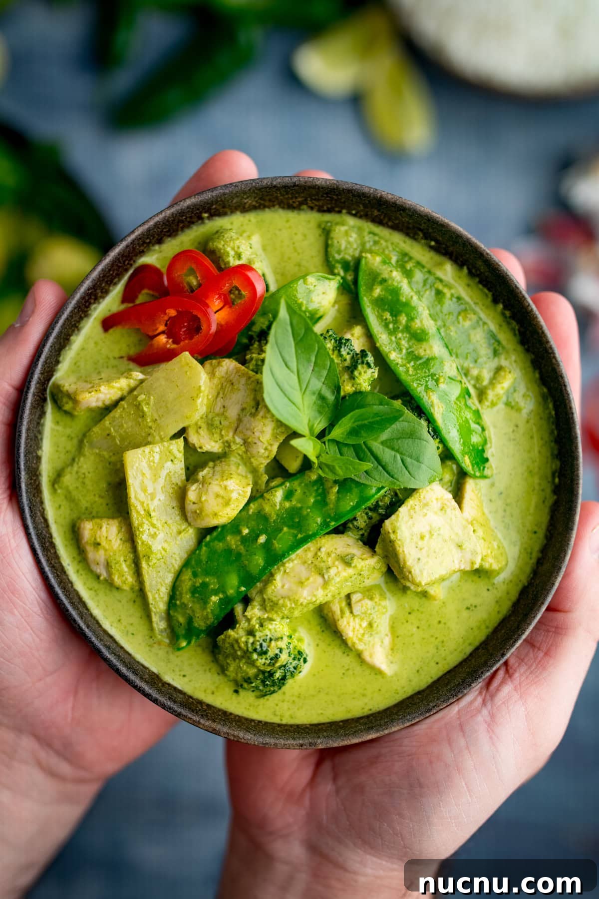 Authentic Thai Green Chicken Curry Recipe (Step-by-Step) 2 A bowl of Thai Green Curry with chicken, mangetout, broccoli, sliced red chilli, and fresh basil, held in two hands.