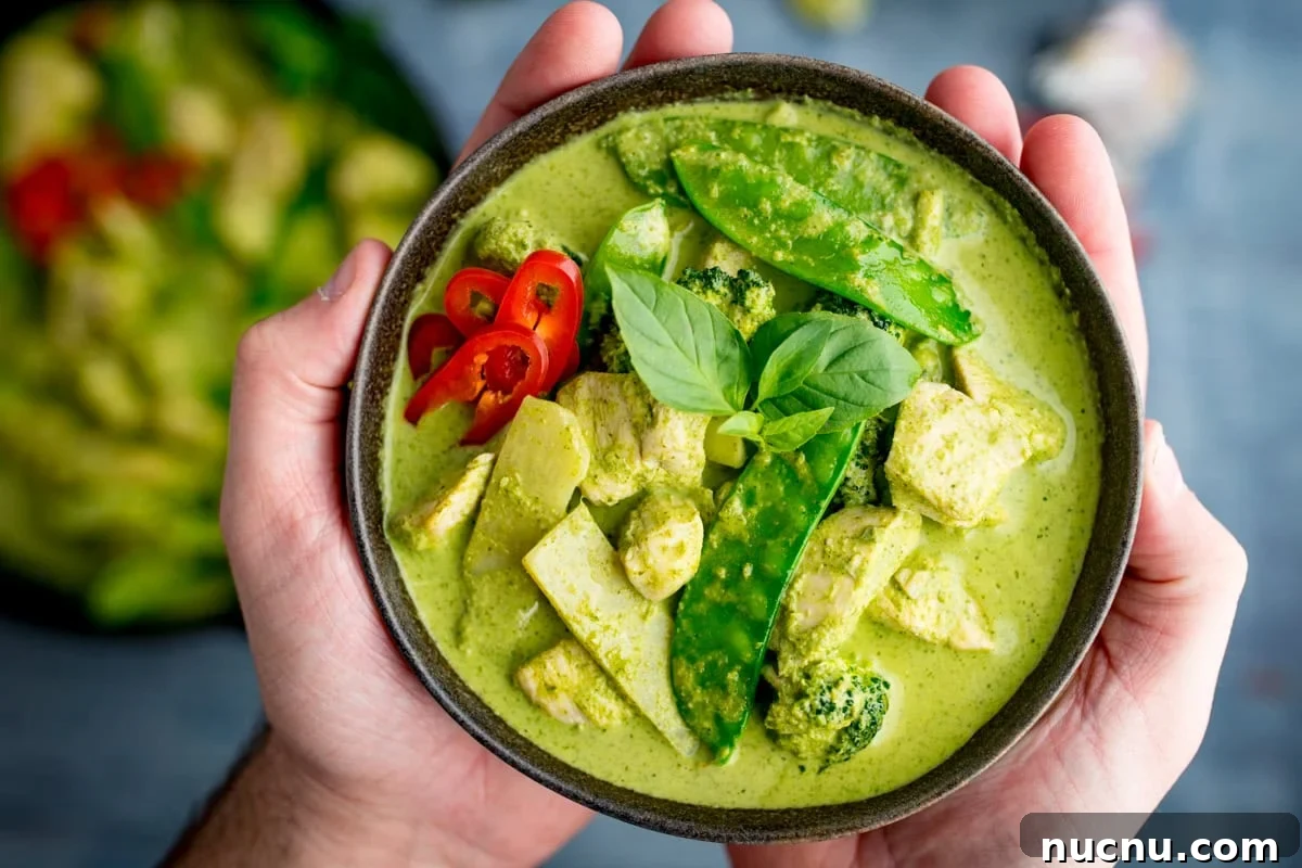 Authentic Thai Green Chicken Curry Recipe (Step-by-Step) 7 A bowl of Thai Green Curry with chicken, mangetout, broccoli, red chilli slices, and fresh basil leaves, held in two hands.