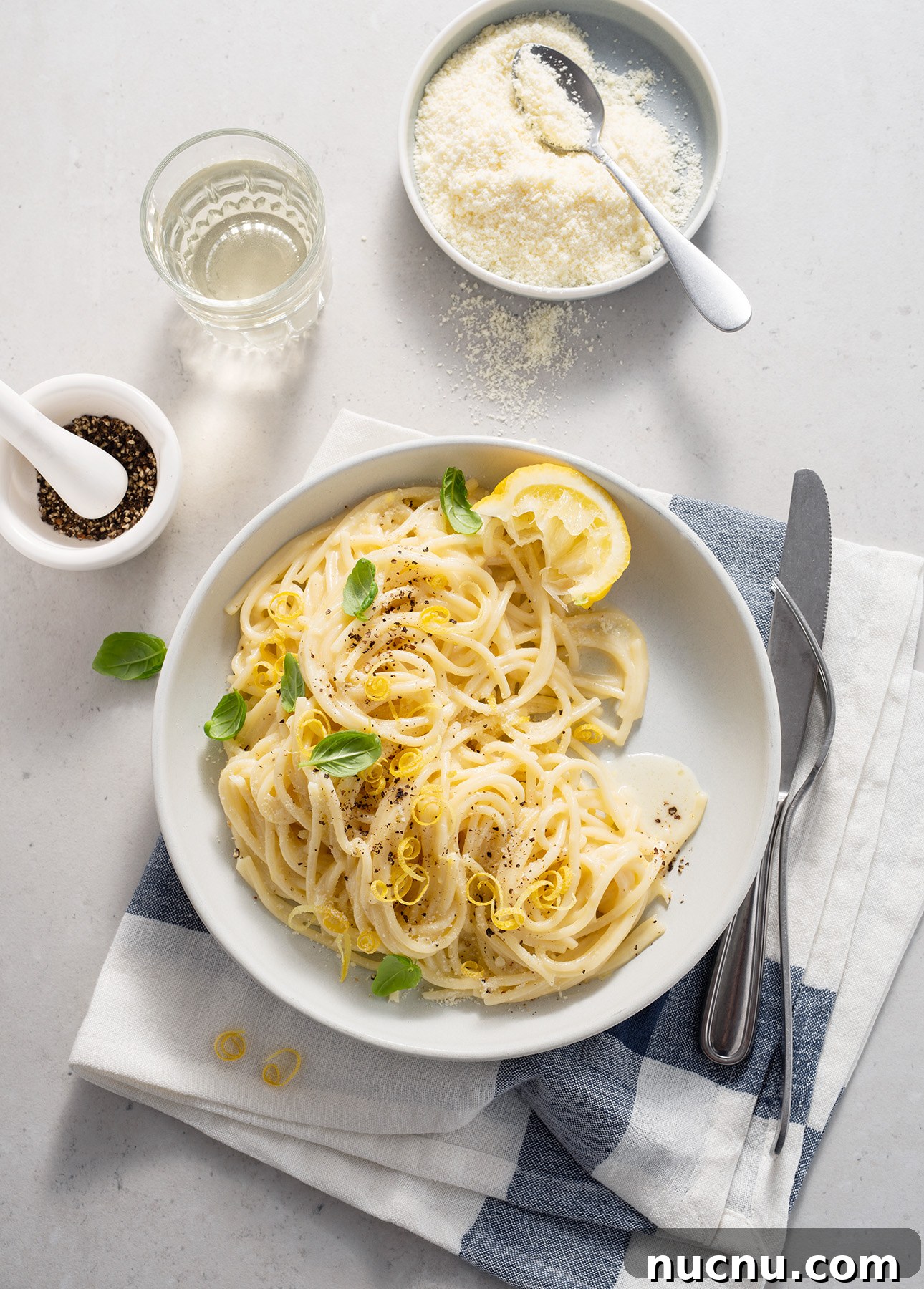 Creamy Lemon Spaghetti Recipe: Zesty, Silky Pasta in 20 Minutes 2 A plate of creamy spaghetti al limone garnished with lemon slices, basil, and black pepper sits on a white napkin with a fork and knife. Nearby are a bowl of grated cheese, a glass of water, and a mortar with peppercorns.