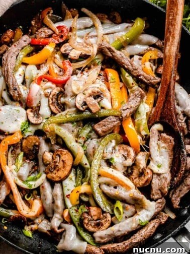 overhead shot of steak strips pepper strips, and mushrooms in a black skillet with brown wooden spoon mixing through the ingredients