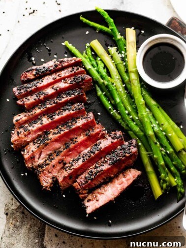 Sliced pan seared sesame crusted tuna steak and served on a black dinner plate with a side of asparagus and soy sauce in a bowl for dipping.