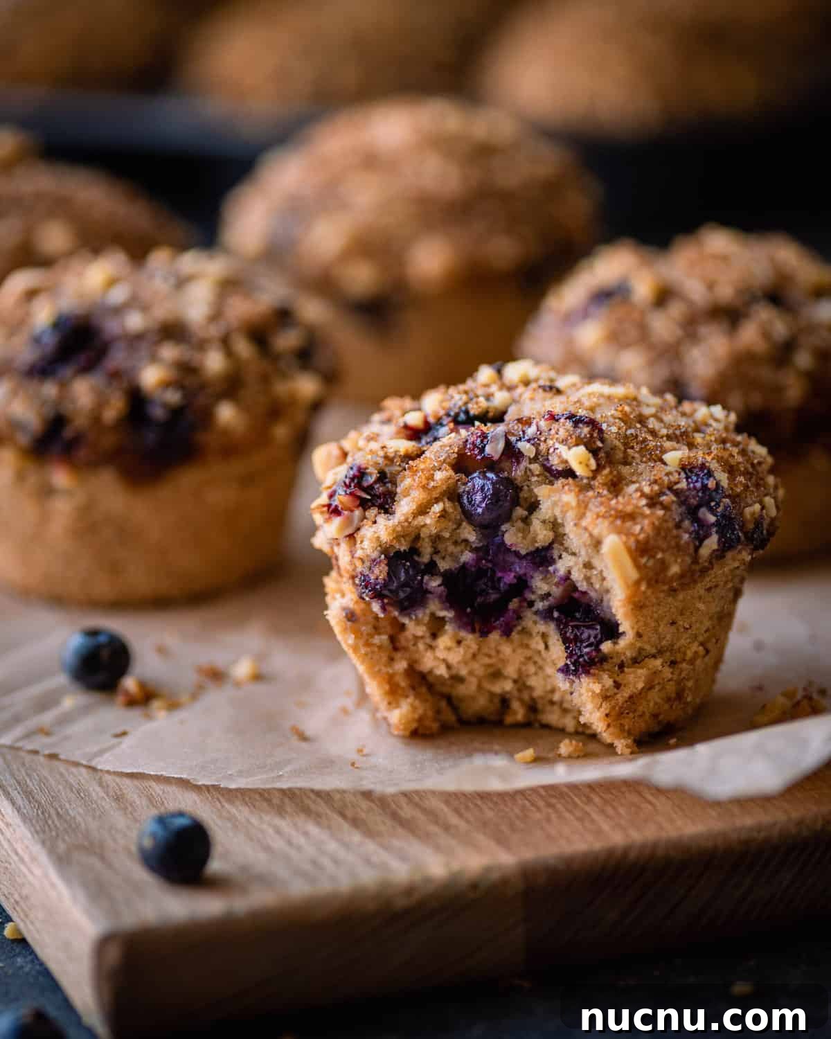 Vegan Blueberry Muffins Recipe: Moist, Fluffy, Simple to Make 2 vegan blueberry muffin with bite shot on parchment paper with other muffins in background.