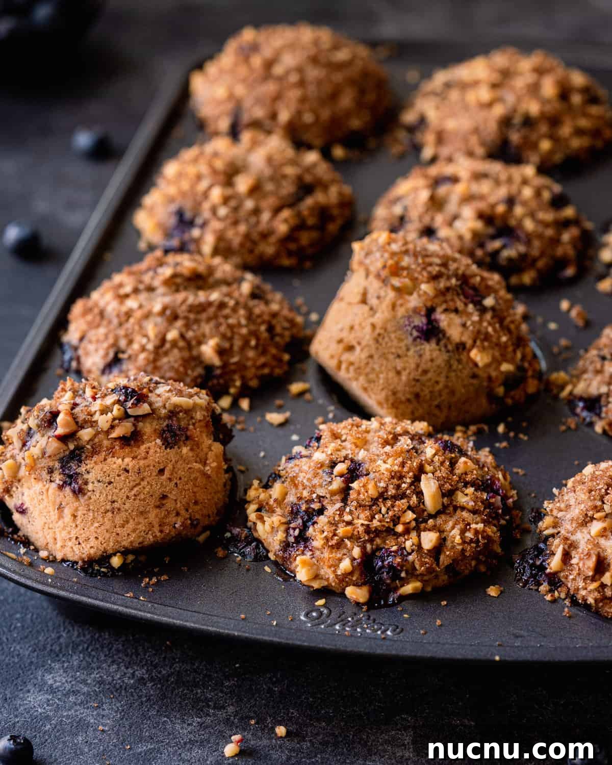 Vegan Blueberry Muffins Recipe: Moist, Fluffy, Simple to Make 3 blueberry muffins with crumble topping in muffin tin, two muffins spilling out.