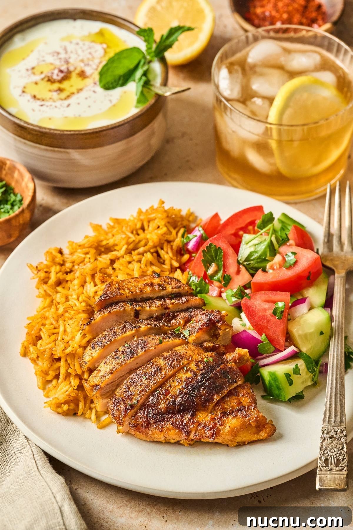 One-Pan Shawarma Chicken and Rice Skillet Recipe 12 A plate with sliced grilled chicken, spiced rice, and a cucumber-tomato salad, with iced tea and dip on the side.