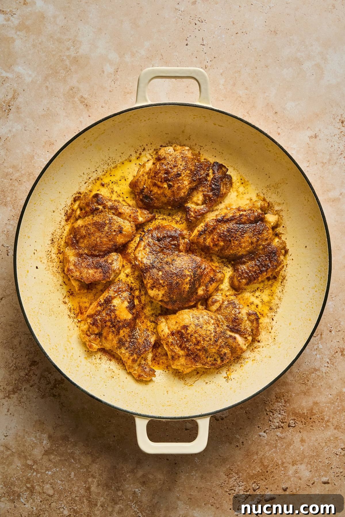One-Pan Shawarma Chicken and Rice Skillet Recipe 15 Golden-brown seasoned chicken thighs cooking in a large cream-colored pan on a textured surface.