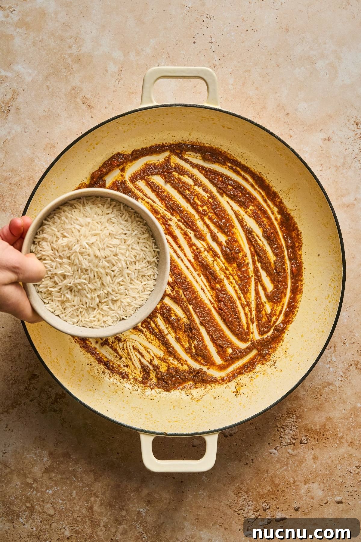 One-Pan Shawarma Chicken and Rice Skillet Recipe 16 A hand pours uncooked rice from a bowl into a pot with a spiced sauce on the bottom.