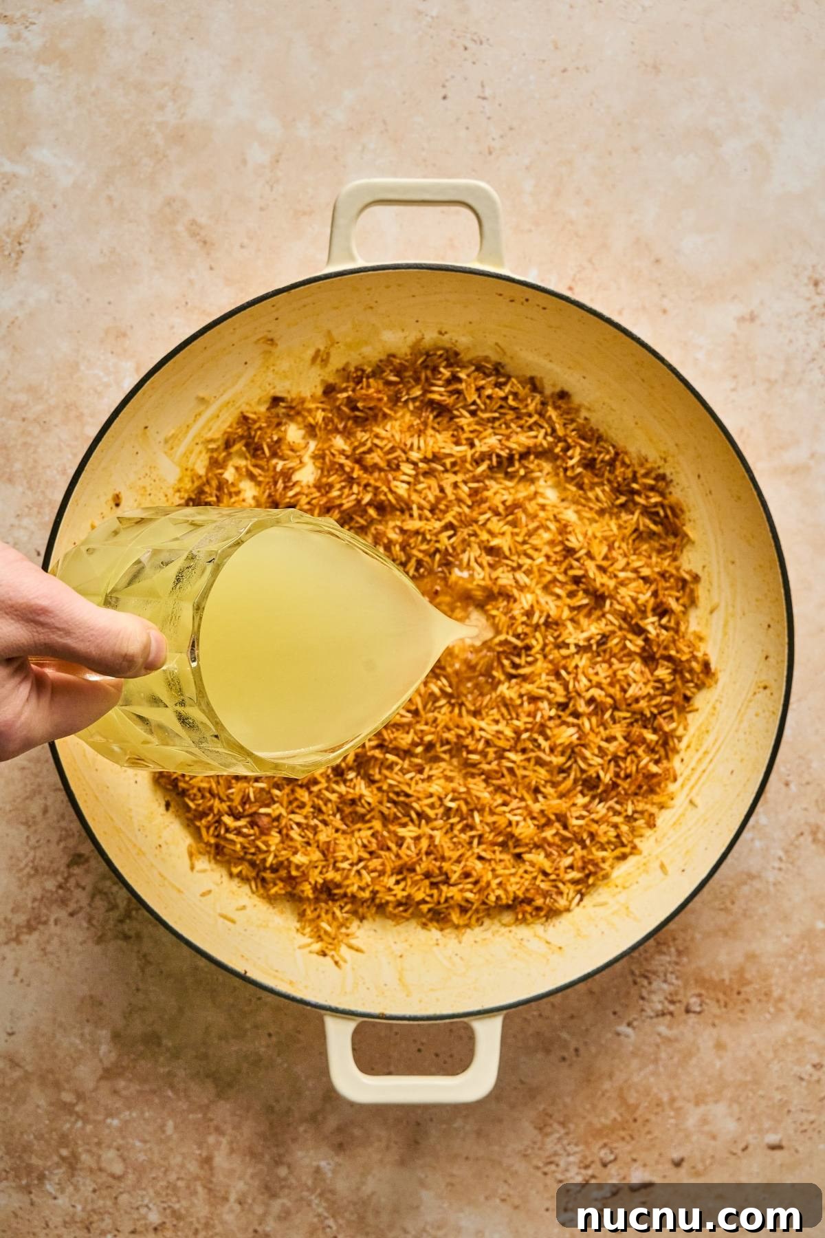One-Pan Shawarma Chicken and Rice Skillet Recipe 17 A hand pours broth from a measuring cup into a pot of toasted rice on a beige surface.