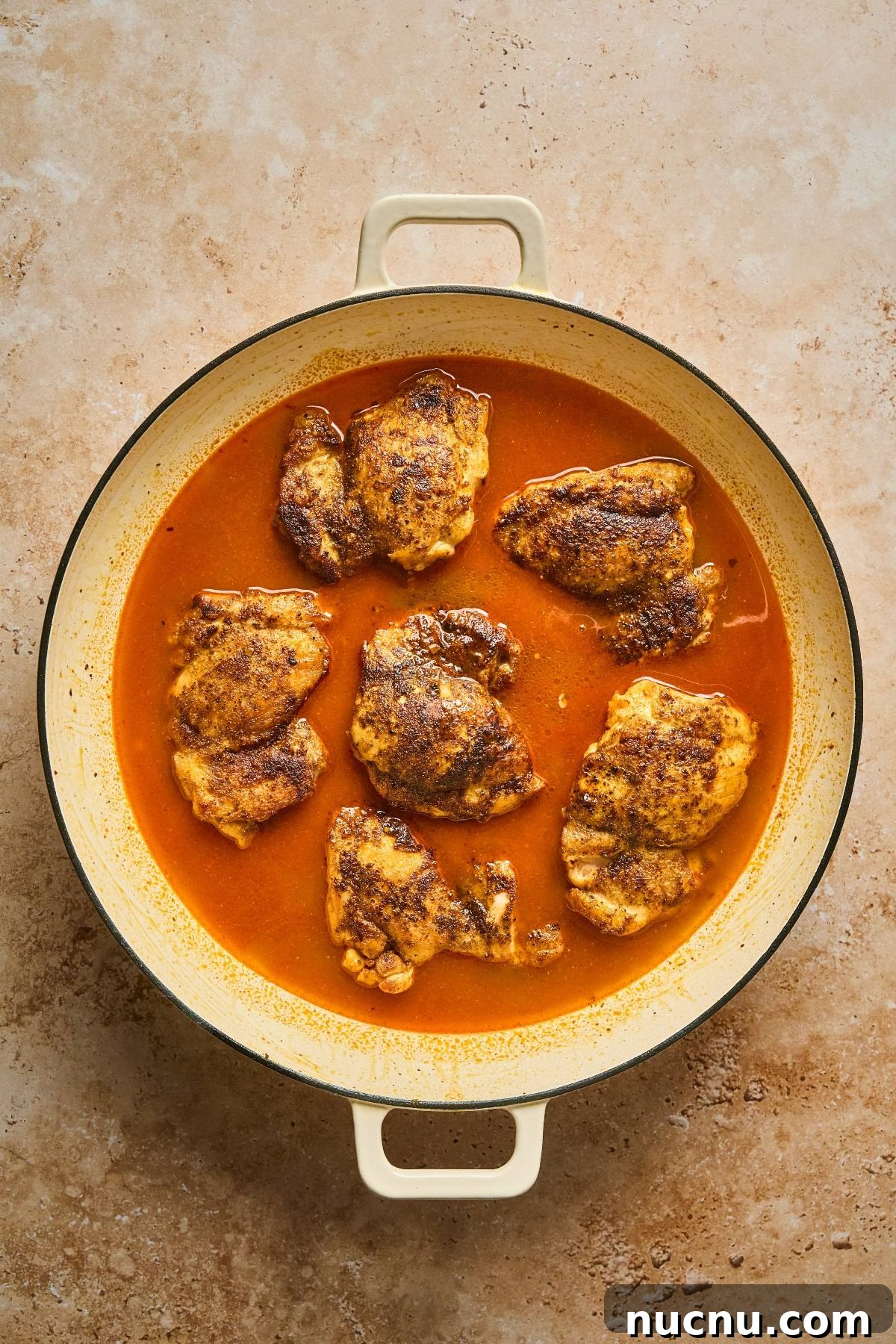 One-Pan Shawarma Chicken and Rice Skillet Recipe 18 Chicken thighs simmer in a round pan with rich red sauce on a tan textured surface.