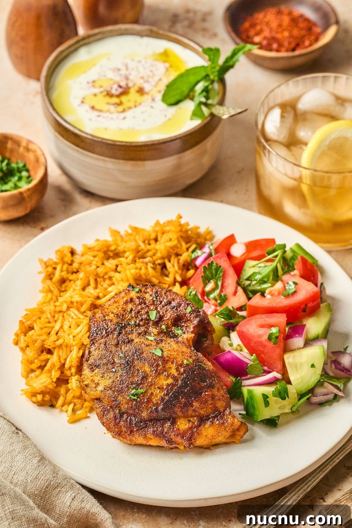 One-Pan Shawarma Chicken and Rice Skillet Recipe 4 Plate with spiced chicken, yellow rice, cucumber-tomato salad; yogurt dip and iced tea in the background.