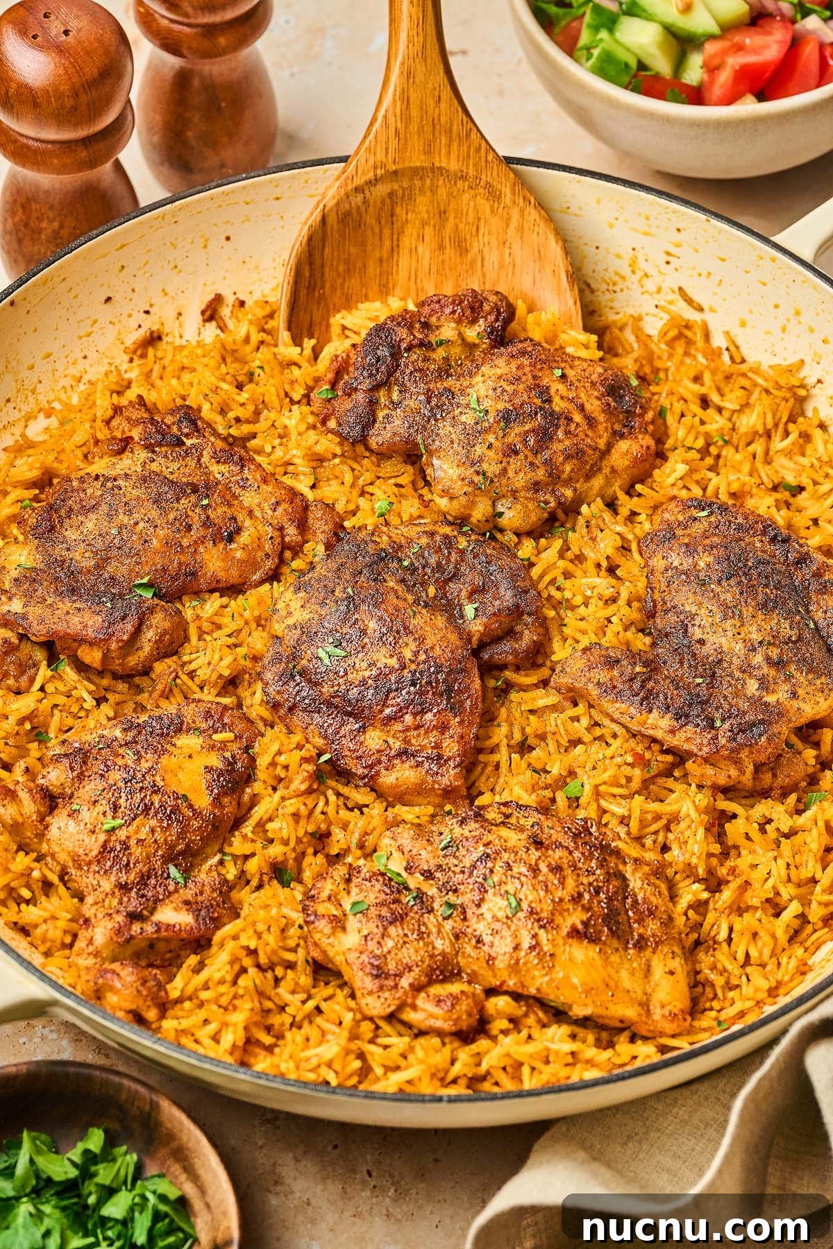 One-Pan Shawarma Chicken and Rice Skillet Recipe 5 A Shawarma Chicken and Rice Skillet features seasoned chicken thighs on yellow rice, with a wooden spoon and a bowl of salad nearby.