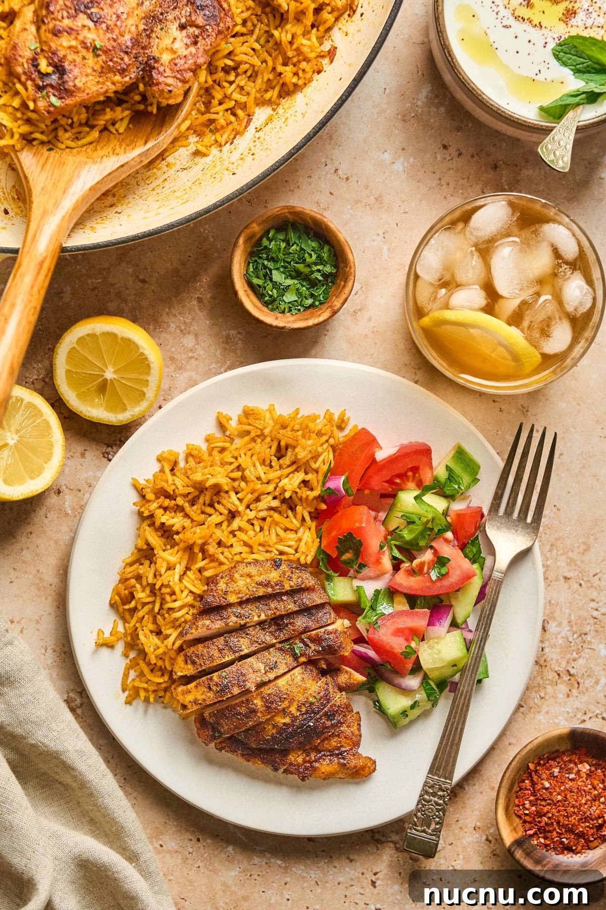 One-Pan Shawarma Chicken and Rice Skillet Recipe 7 A plate with spiced rice, grilled chicken, salad, and a fork, with iced tea, lemon, and herbs nearby.