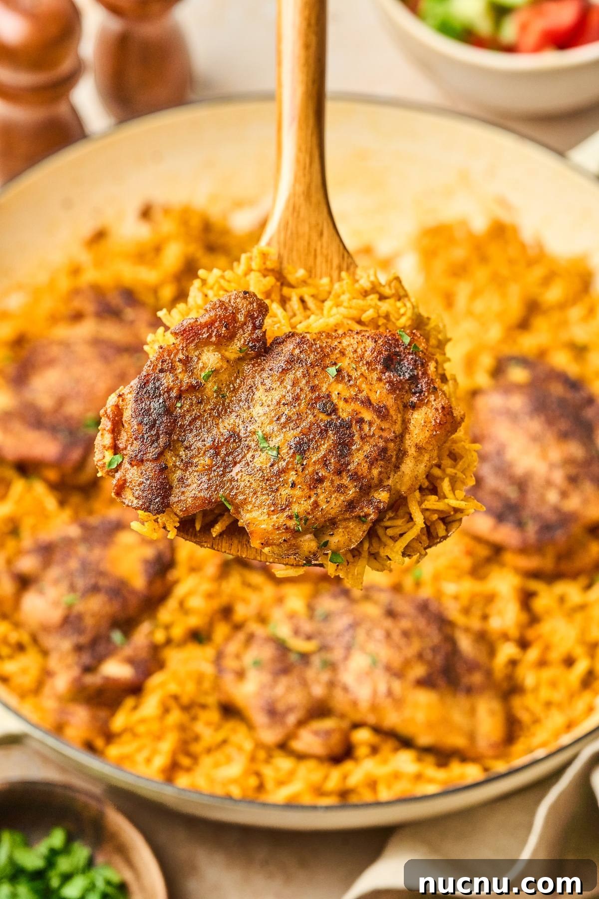 One-Pan Shawarma Chicken and Rice Skillet Recipe 10 A wooden spoon holds seasoned chicken thigh over a pan of yellow rice and more chicken thighs.