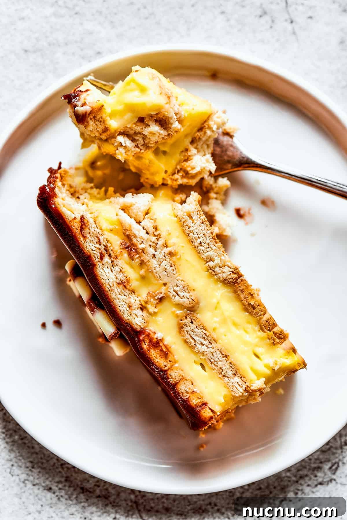 Chocolate eclair cake is loaded with creamy and smooth pudding, decadent chocolate ganache, and tender cookies, and it's so easy to make. Overhead view of a piece of chocolate eclair cake on a plate, with a bit of it broken off with a fork.