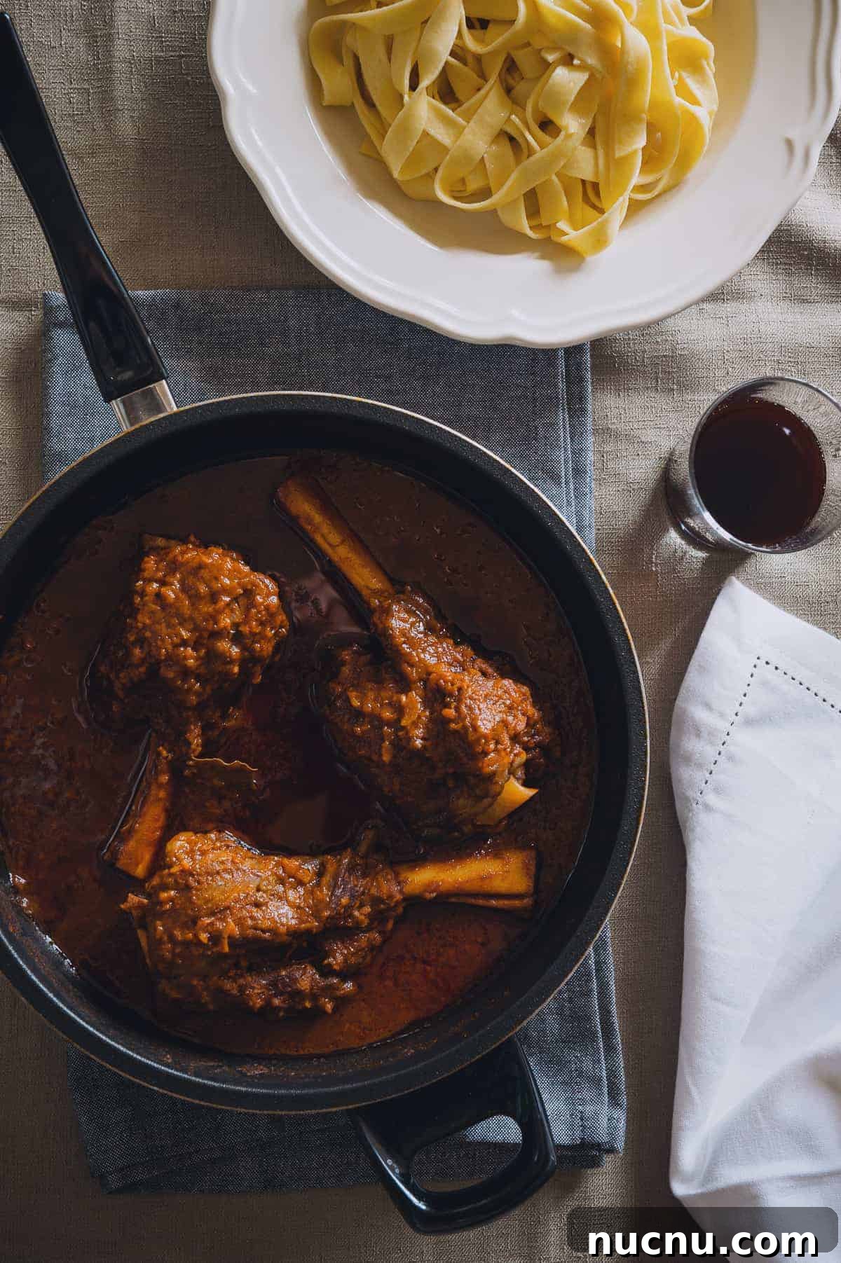 top down view of a pan containing three braised lamb shanks and a bowl of noodles