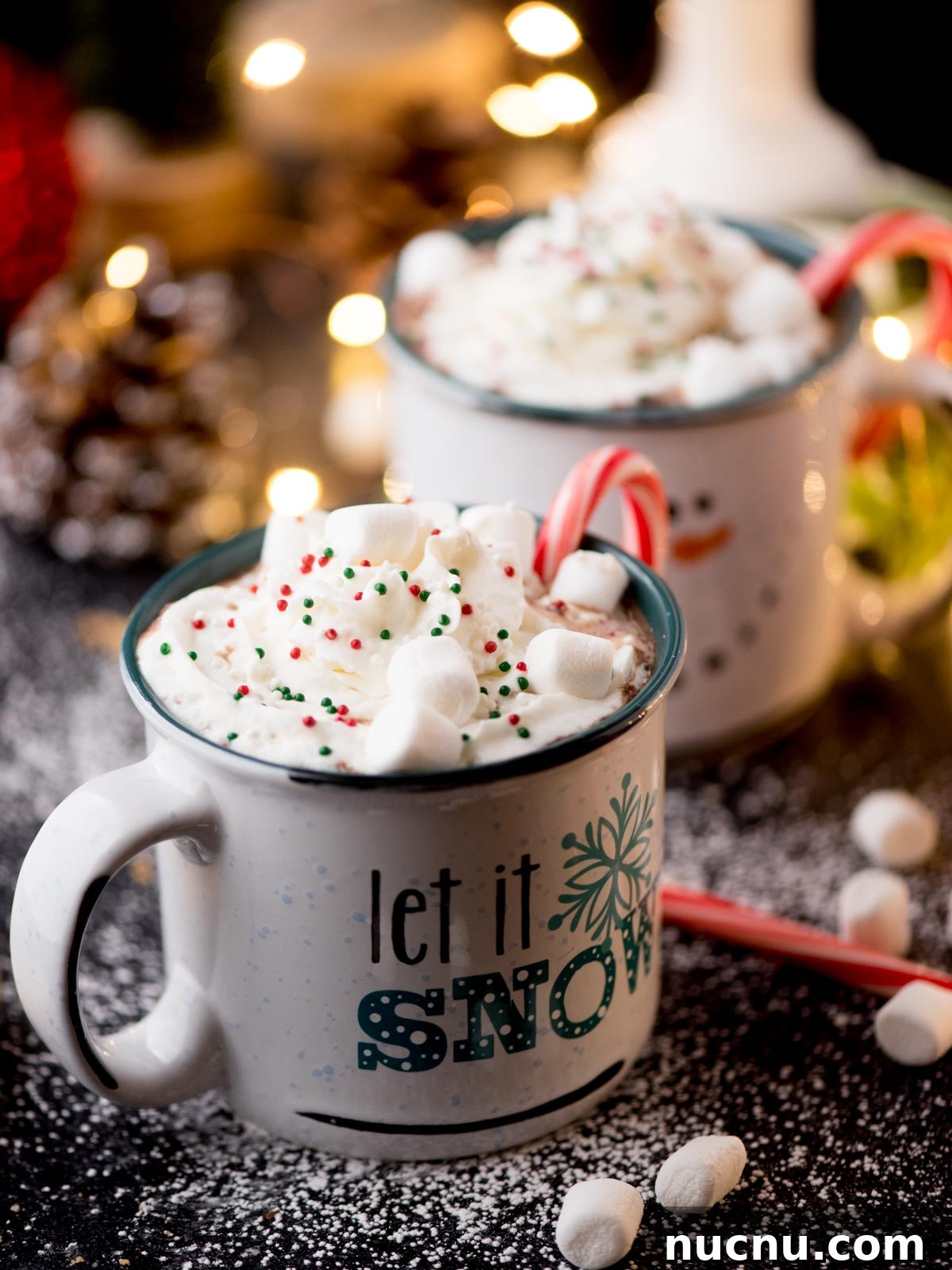 Creamy Crockpot Hot Chocolate Recipe for Cozy Nights 2 Two mugs of hot chocolate topped with whipped cream, colorful sprinkles, marshmallows, and candy canes. The front mug features the text let it snow. A festive background with fairy lights and snow-like powder completes the scene.