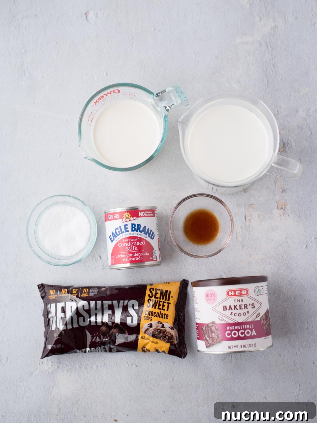 Creamy Crockpot Hot Chocolate Recipe for Cozy Nights 3 Ingredients for a dessert are laid out on a light surface: a bag of semi-sweet chocolate chips, a can of sweetened condensed milk, a small bowl of sugar, a glass of vanilla extract, a carton of heavy cream, and a box of unsweetened cocoa.