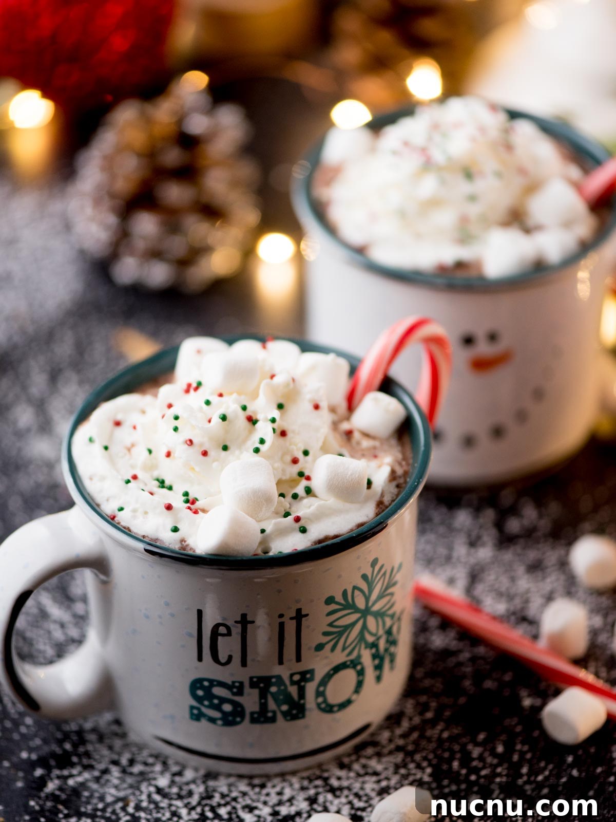 Creamy Crockpot Hot Chocolate Recipe for Cozy Nights 4 Two festive mugs of hot chocolate topped with whipped cream, sprinkles, and marshmallows. One mug has a candy cane and Let it Snow printed on it. The scene is sprinkled with powdered sugar, and there are twinkling lights and a pinecone in the background.