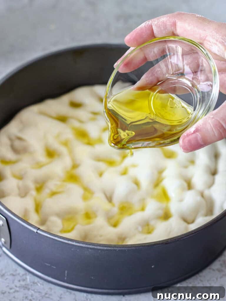 Tomato Focaccia Recipe with Step-by-Step Video Tutorial 6 Drizzling oil on dimpled focaccia dough in a round dark metal cake pan.