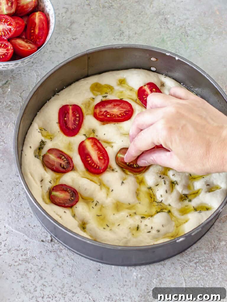 Tomato Focaccia Recipe with Step-by-Step Video Tutorial 7 Placing halved cherry tomatoes on focaccia dough in a round pan. Gray background.
