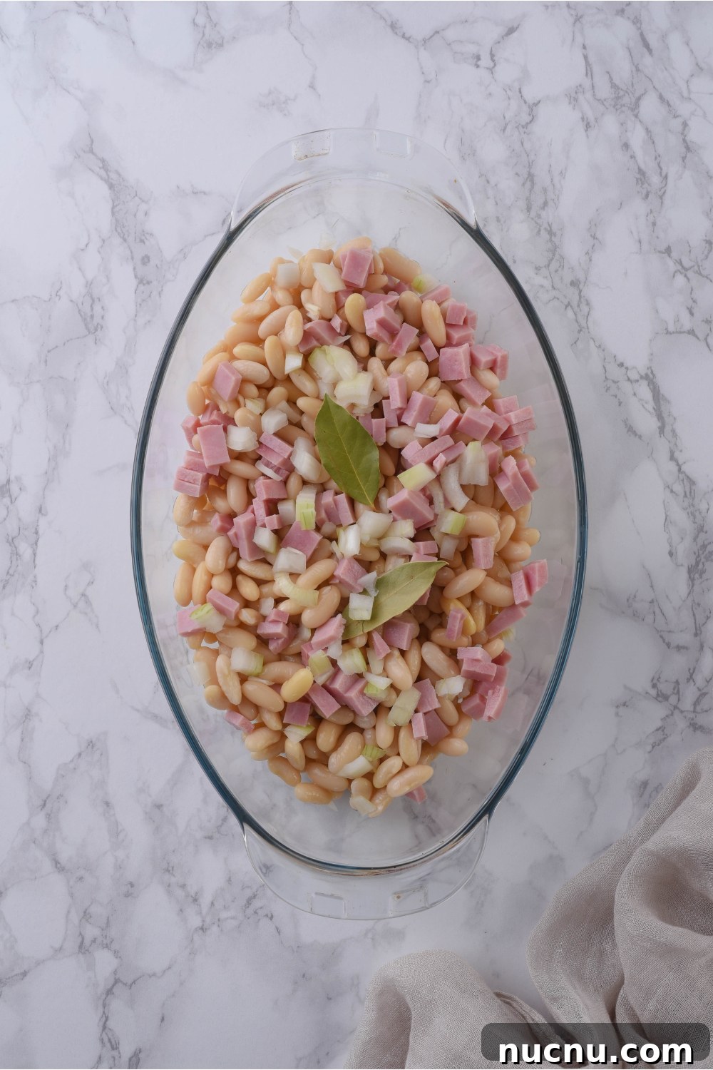 Copycat Grandma Brown's Baked Beans Recipe You’ll Love 7 A clear baking dish filled with baked beans, diced onion, cubed ham, and two bay leaves.