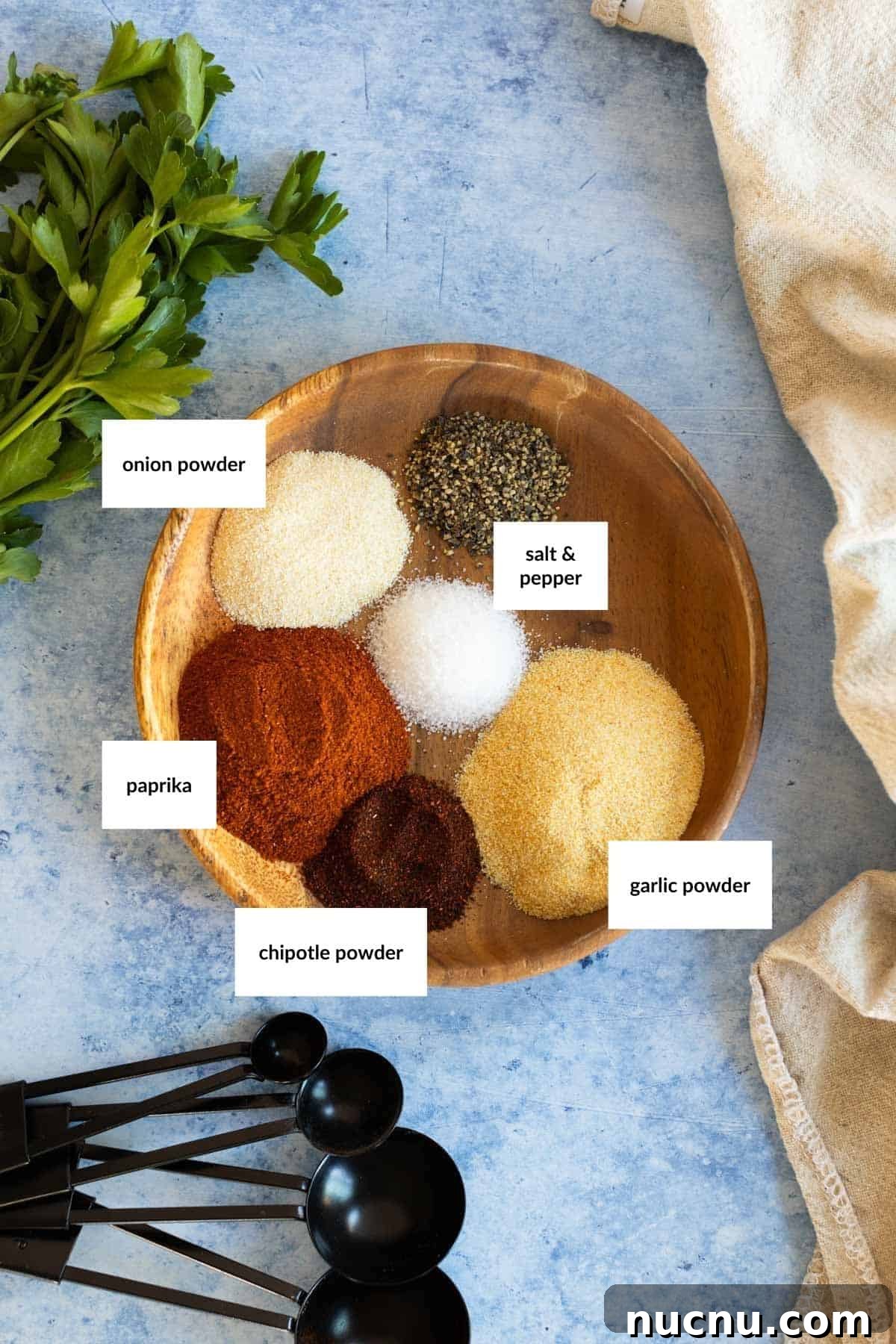 Crispy Dry-Rub Wings Recipe for Perfectly Spiced Chicken 3 A wooden bowl containing paprika, chipotle powder, onion powder, garlic powder, salt, pepper, surrounded by parsley, measuring spoons, and a beige cloth on a blue surface.