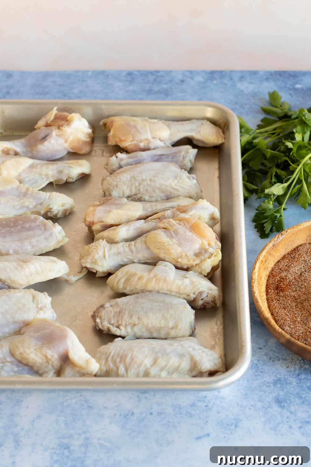 Crispy Dry-Rub Wings Recipe for Perfectly Spiced Chicken 7 Raw chicken wings on a baking tray beside a bowl of seasoning and parsley on a kitchen counter.