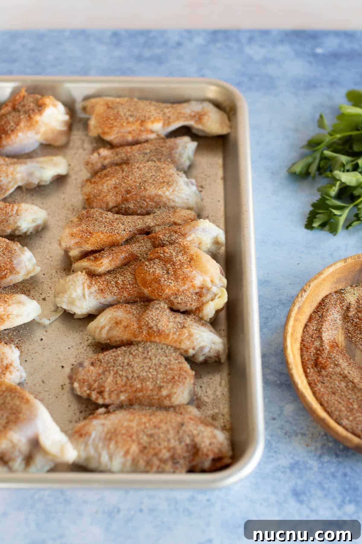 Crispy Dry-Rub Wings Recipe for Perfectly Spiced Chicken 8 Raw chicken wings seasoned with dry rub on a baking sheet, alongside fresh parsley and a bowl of additional spices.