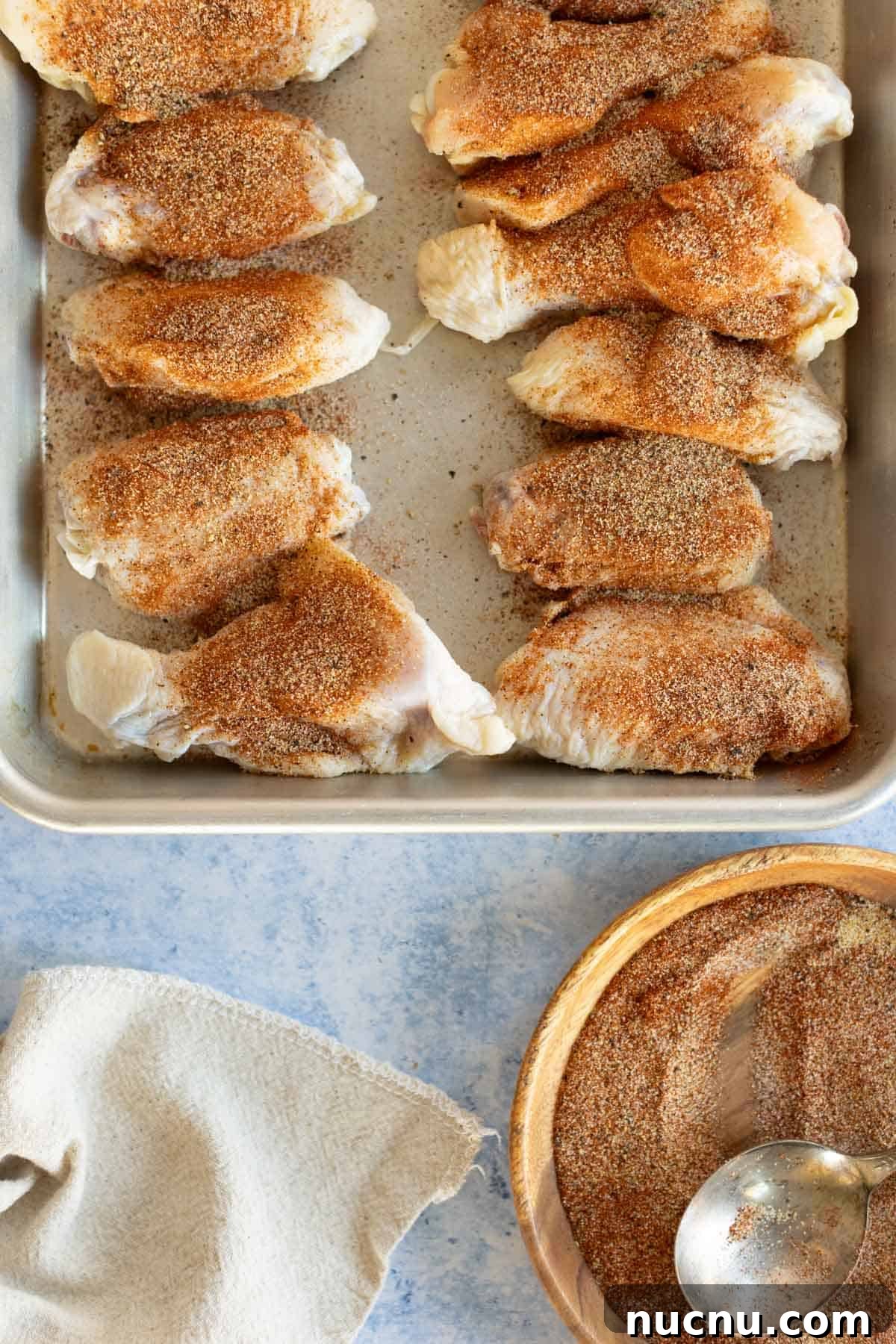 Crispy Dry-Rub Wings Recipe for Perfectly Spiced Chicken 9 Raw chicken breasts seasoned with spices on a metal baking tray, next to a wooden bowl with additional spices and a spoon.