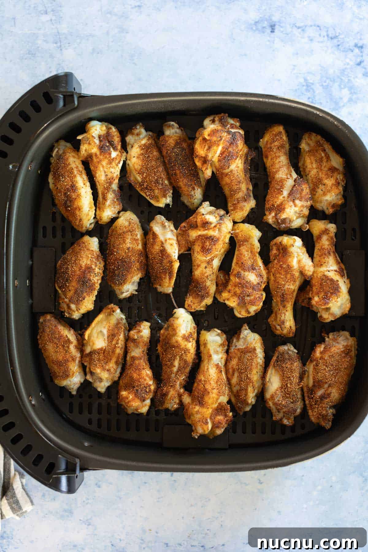 Crispy Dry-Rub Wings Recipe for Perfectly Spiced Chicken 10 Golden-brown drumsticks seasoned with a dry rub arranged in a black air fryer basket on a light blue surface.