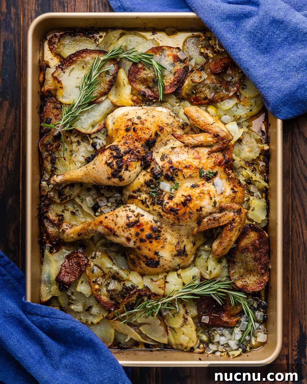 Spatchcock chicken and potatoes on baking sheet with rosemary garnish and blue napkins.