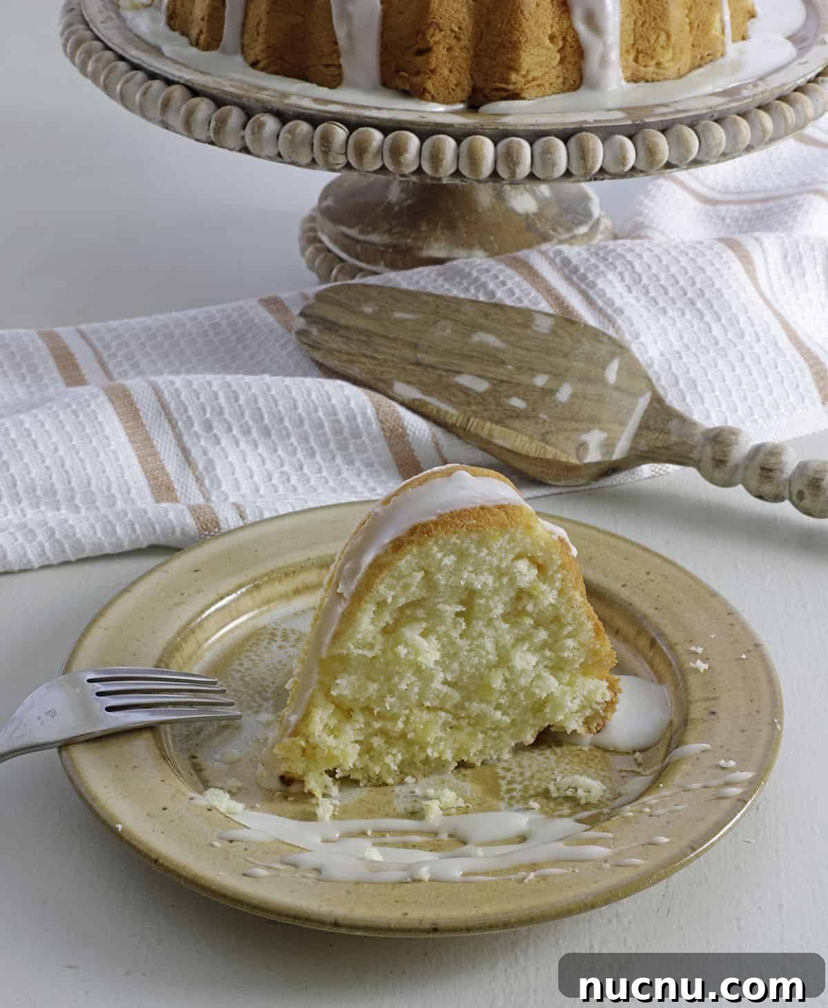 Slice of glazed cake on brown stone plate with fork on the side and baked cake in the background.