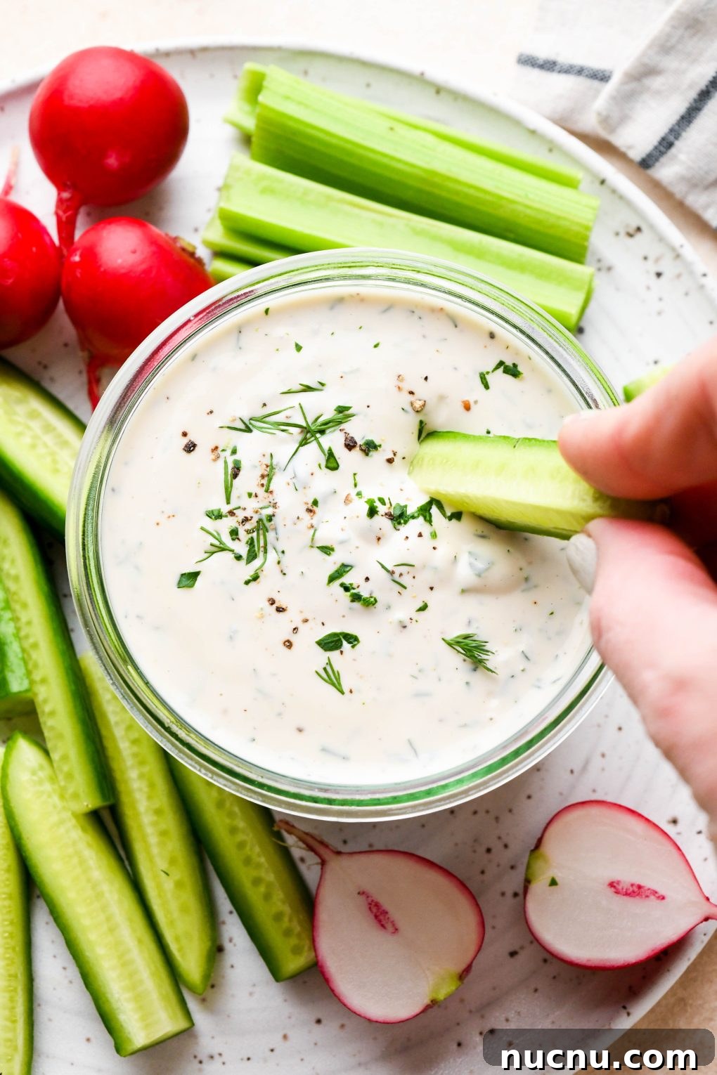 Creamy Raw Cashew Vegan Ranch Dressing Recipe 2 Vegan ranch dressing in a small glass jar with a hand dipping a cucumber slice surrounded by fresh cut veggies.