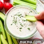 Vegan ranch dressing in a small glass jar with a hand dipping a cucumber slice surrounded by fresh cut veggies.