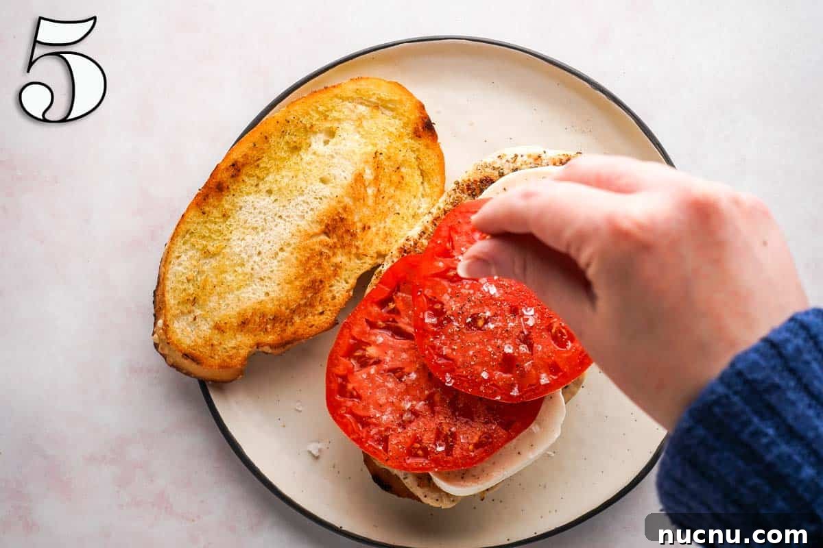 Caprese Chicken Sandwich with Fresh Mozzarella & Basil 8 hand sprinkling salt on top of a tomato with a slice of bread on the side