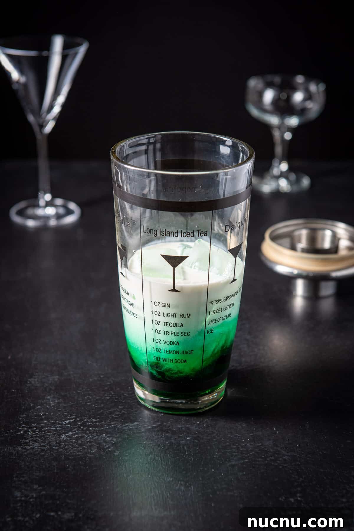 A shaker filled with a green liqueur, and cream with two glasses in the back