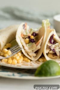 three fish tacos with spicy slaw on a plate with a fork, corn and lime wedge